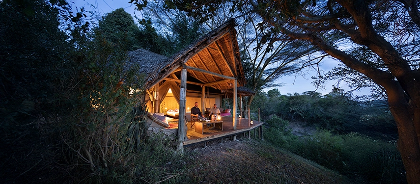 Evening view of a guest relaxing in a candlelit tented suite nestled in the forest at Basecamp Masai Mara, with warm ambient lighting.