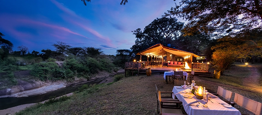 Riverside tented lounge and dining area at Basecamp Masai Mara, set for dinner under the stars with lanterns glowing at dusk.