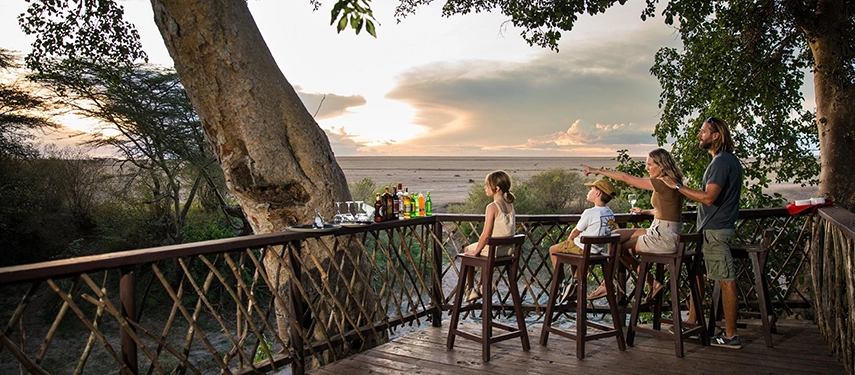 Family enjoying sunset drinks on a wooden deck overlooking the plains at Basecamp Masai Mara, with a scenic view of the savannah horizon.