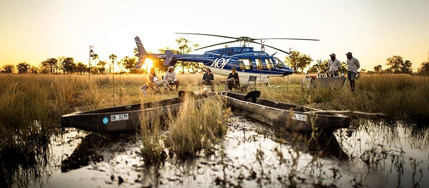 A helicopter transfer lands beside traditional mokoro canoes, highlighting the seamless blend of air and water travel in the Okavango Delta.