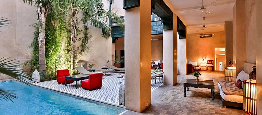 Courtyard pool at Dar Bensouda surrounded by palm trees, red chairs, and Moroccan tiled flooring.