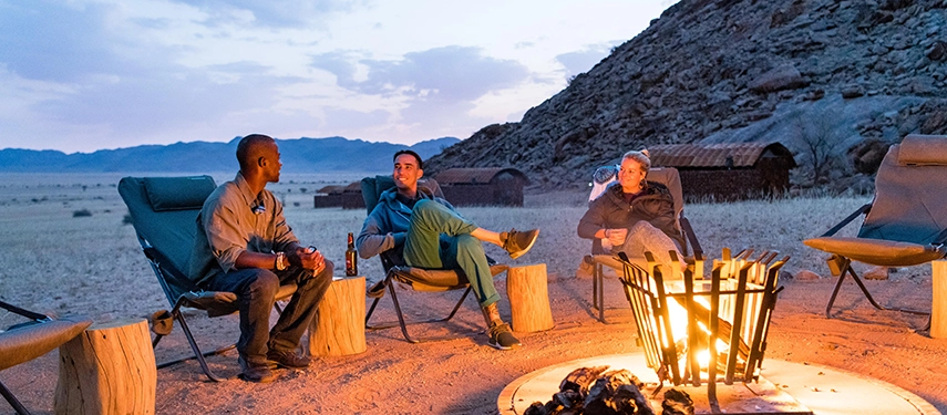 Guests relaxing around a glowing campfire at Camp Sossus with desert plains and rocky hills in the background during twilight.