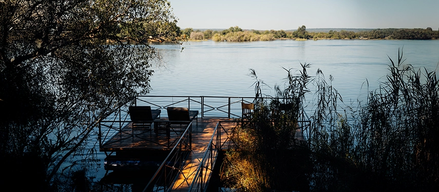 A wooden riverside platform at Chundukwa River Lodge provides peaceful seating above the flowing waters of the Zambezi.