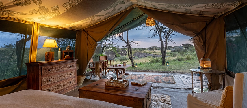 A luxury safari tent interior at dusk, open to the Mara bush beyond, with warm lamplight, a carved chest and a woven pendant light casting a soft glow.