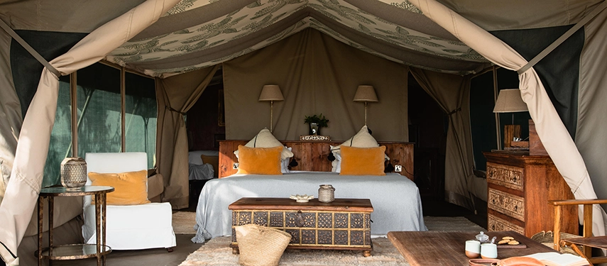 The interior of a classic safari tent at Hemingways River Camp Mara, featuring a king bed with ochre cushions, carved wooden furniture and a patterned canvas ceiling.