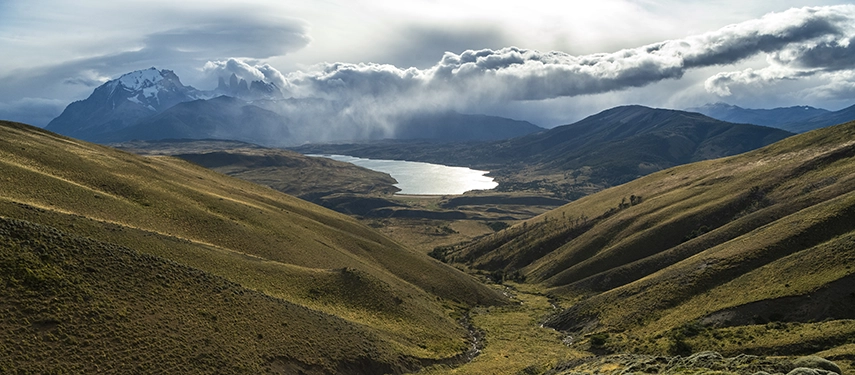 Golden hills fall away to a shimmering lake as storm clouds sweep across the Patagonian mountains beyond.