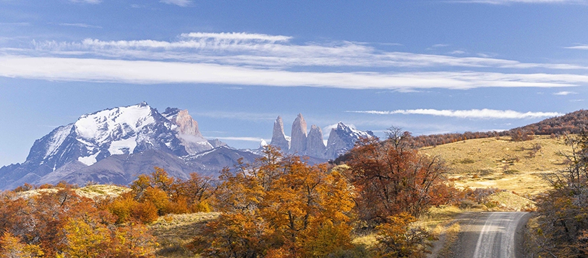 A winding gravel road curves through autumn-coloured hills towards the snow-capped peaks and granite towers of Torres del Paine.