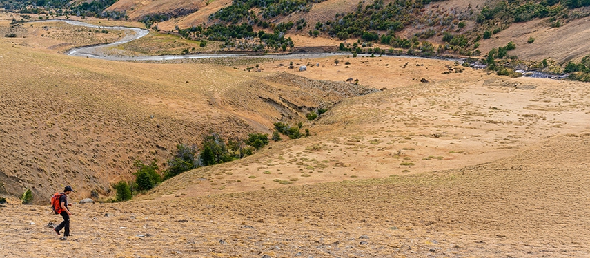 A lone hiker crosses ochre hills that fall away to a winding Patagonian river below.