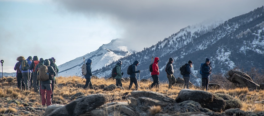 A line of hikers crosses a windswept Patagonian slope, framed by rugged mountains dusted with early winter snow.
