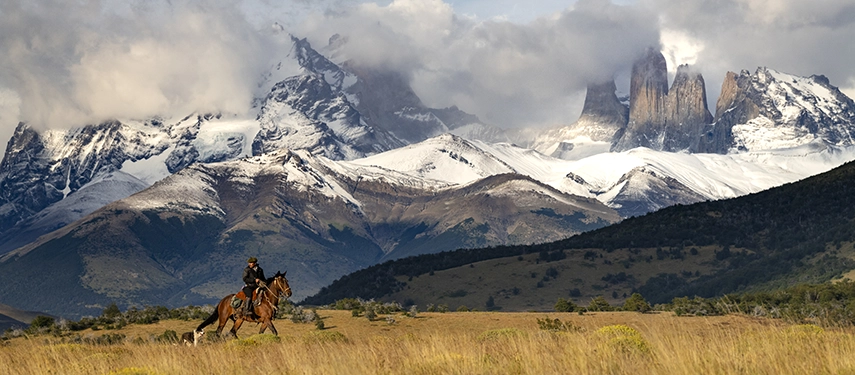 A lone rider moves across open grasslands with the dramatic snow-capped Paine massif towering in the background.