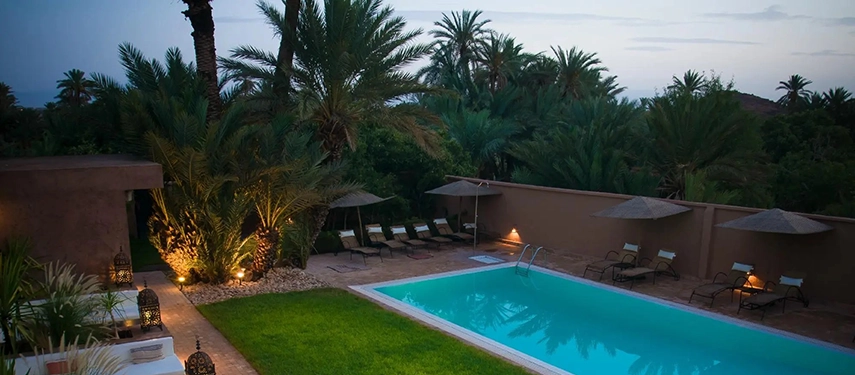 Palm-fringed swimming pool at dusk with sun loungers set against the garden wall.