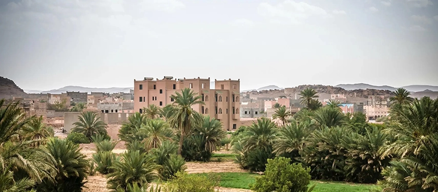 Exterior view of Dar Chamaa rising above palm groves and village rooftops with rocky hills beyond.