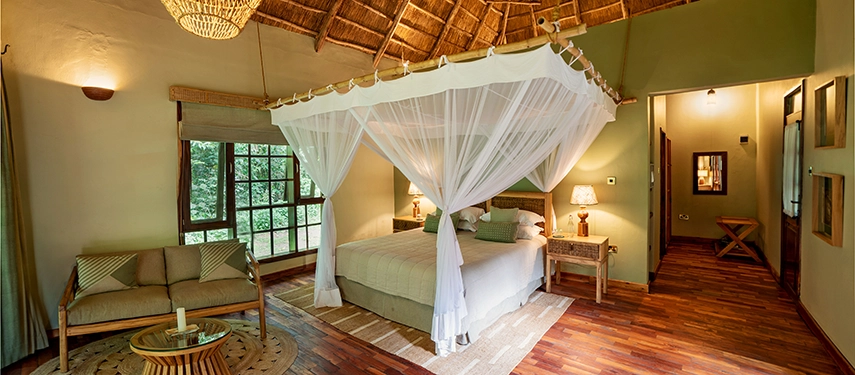 Traditional thatched-roof double cottage at Primate Lodge Kibale with mosquito-draped bed and seating area.