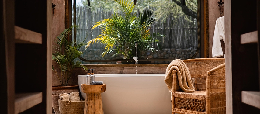 A serene en-suite bathroom in the Family Tent at Hemingways River Camp Mara, with a freestanding white bath, tropical plants and natural light filtering through a garden window.
