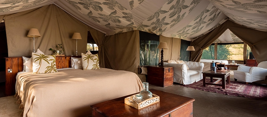 A spacious Family Tent bedroom at Hemingways River Camp Mara, with a double bed, palm-print cushions, polished wood furnishings and a lounge area beyond.
