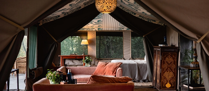 The shared lounge area of the Family Tent at Hemingways River Camp Mara, with a deep coral sofa, inlaid wooden cabinet and soft lamplight filtering through canvas walls.