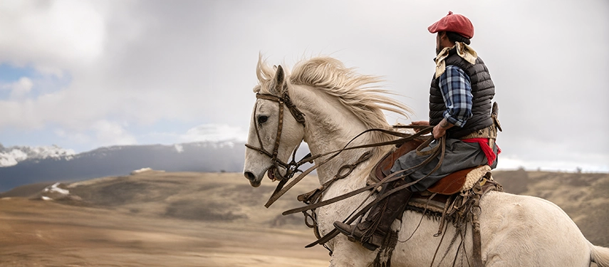 A gaucho on a white horse rides across open Patagonian plains, framed by distant mountains and soft winter light.