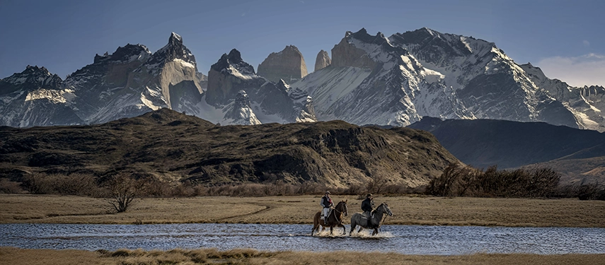 Two riders guide their horses through a shallow river with the jagged, snow-capped Paine massif rising sharply behind them.