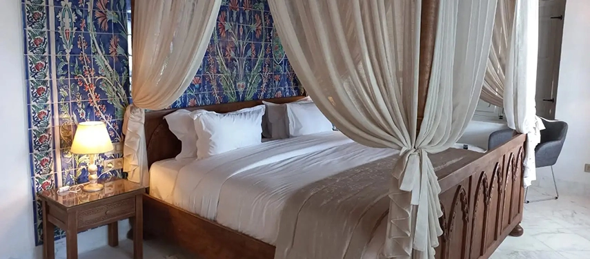 Traditional bedroom with a carved wooden four-poster bed, sheer drapes, and patterned tiled wall.