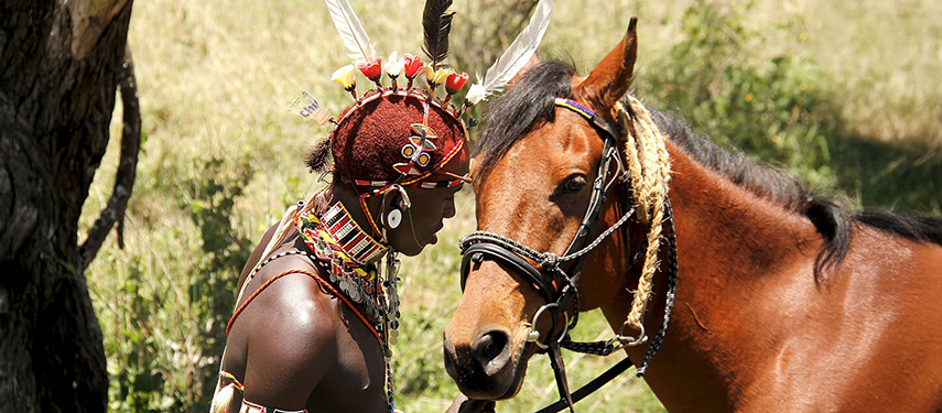 A Samburu man dressed in ceremonial attire gently touches foreheads with a brown horse, highlighting their bond.