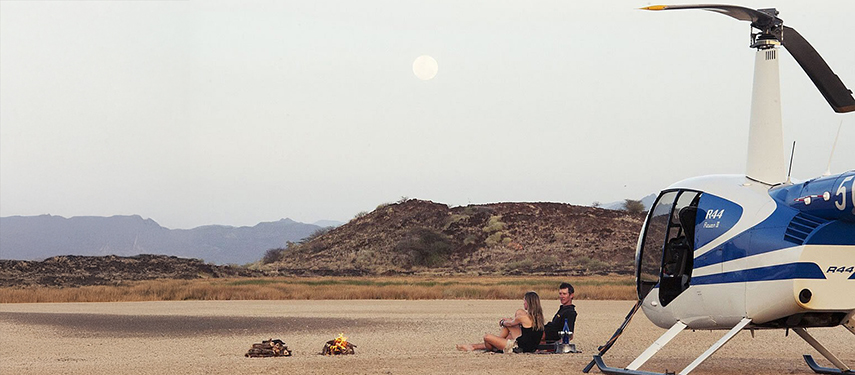 A couple relaxes beside a fire near a helicopter at dusk, surrounded by moonlit plains and distant rocky hills.