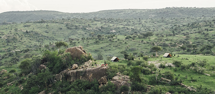 Two canvas tents perched atop a rocky outcrop overlooking a vast green valley, offering a panoramic view of the northern Kenyan wilderness.
