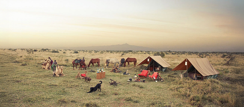 A remote fly camp set up on open plains at dusk, with camels, horses, red chairs, and Samburu guides preparing for the next leg of the journey.