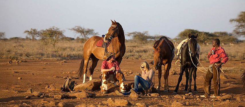 Two travellers and a guide rest beside their saddled horses in a dry, open landscape, taking a break during a horseback safari.