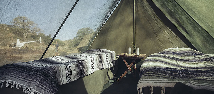 Inside view of a twin safari tent with striped blankets and canvas walls, looking out onto a light aircraft parked nearby.