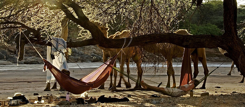 A hammock strung between trees provides a shady retreat, with camels and Samburu guides in the background along a dry riverbed.