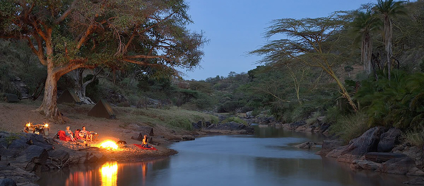 A group enjoys a riverside campfire under a large tree, with canvas tents set up near the water in a remote wilderness setting.