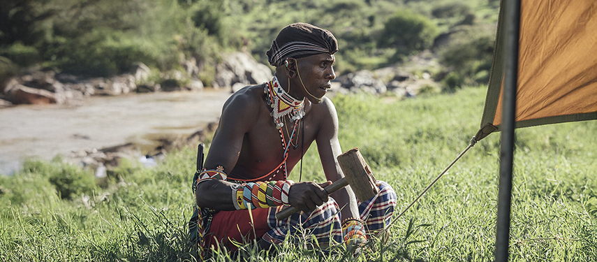 A Samburu warrior in traditional dress drives a tent peg into the grass while setting up camp near a tranquil river.