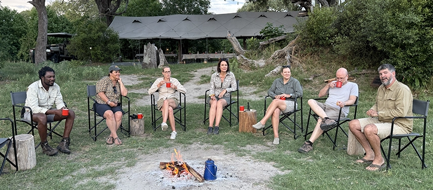 Guests relax around a crackling firepit with mugs in hand, sharing stories during an intimate evening gathering at Karangoma in the Okavango Delta.