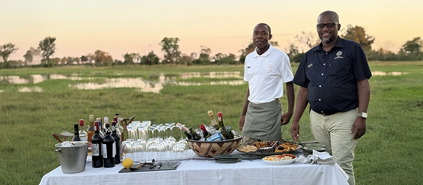 Safari hosts stand beside a beautifully arranged bush dining table, offering drinks and canapés against the open floodplains of Karangoma at sunset.
