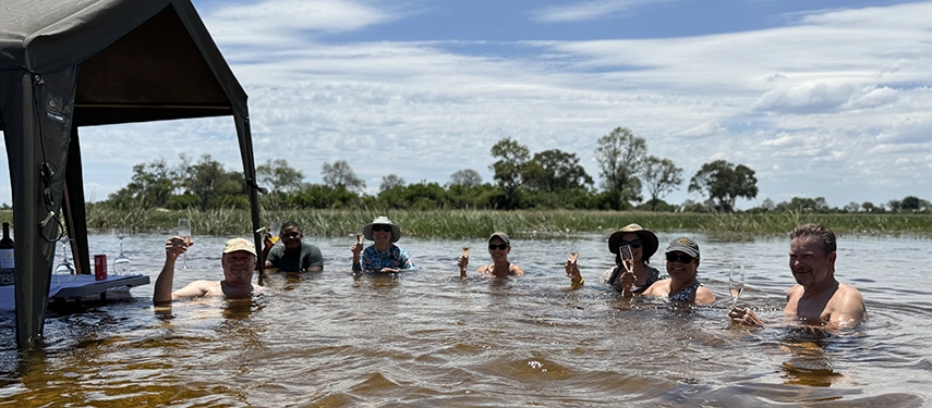 Guests relax in shallow waters with drinks in hand, enjoying a playful safari moment that blends wilderness immersion with light-hearted indulgence.