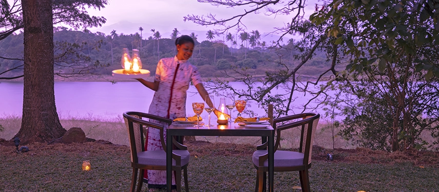 A romantic lakeside setting at Jetwing Kandy Gallery is prepared with candlelight as a staff member serves dinner at dusk.
