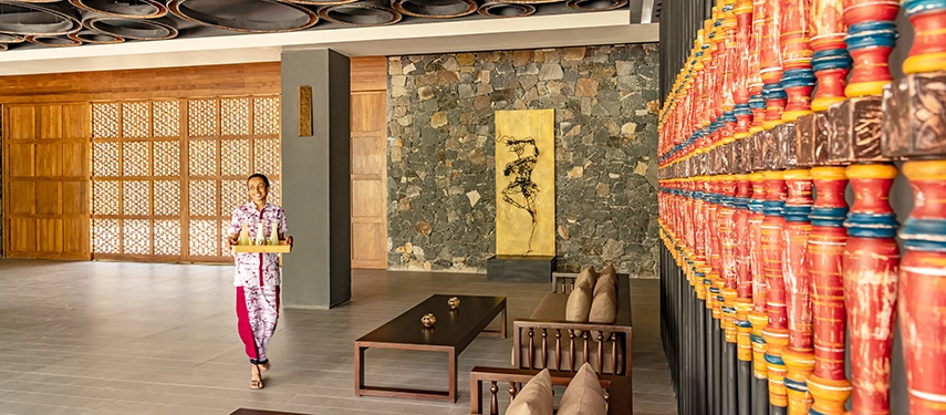 The lobby at Jetwing Kandy Gallery features warm stone walls, vibrant decorative panels and a staff member offering refreshments.