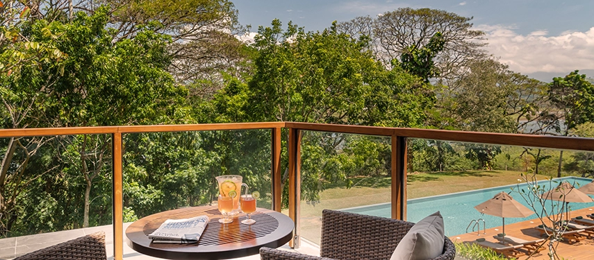 A sunlit balcony at Jetwing Kandy Gallery offers leafy views and a glimpse of the pool below, set with drinks and a relaxed seating area.
