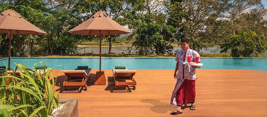A staff member walks past the pool at Jetwing Kandy Gallery carrying refreshments, with sun loungers and treetop views surrounding the serene deck.