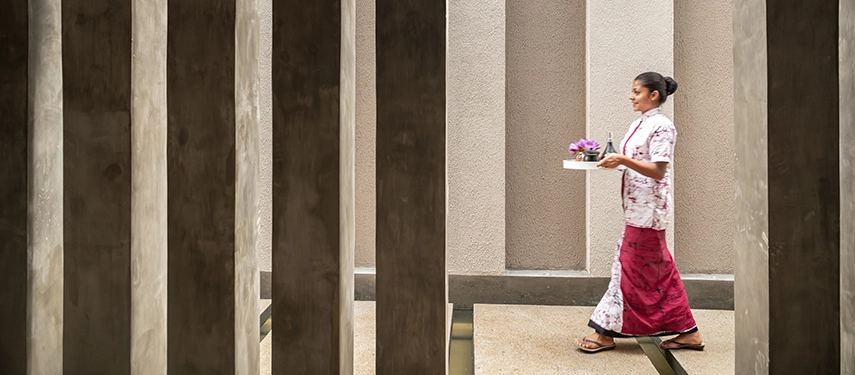 A spa attendant at Jetwing Kandy Gallery walks through a calming corridor carrying fresh flowers on a tray, framed by soft light and textured pillars.