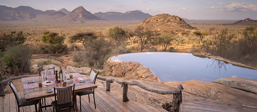 A dining table set on a wooden deck overlooks an infinity pool and sweeping savannah views at Sarara Camp in northern Kenya.