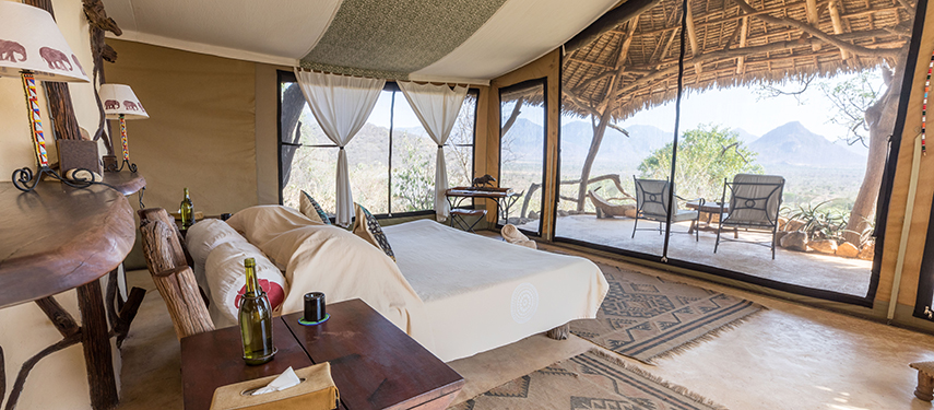 An elegant tented suite at Sarara Camp featuring earthy decor, a large bed, and a shaded verandah with mountain views.