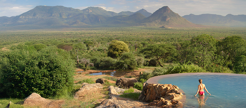 A guest wades into the horizon-edge pool at Sarara Camp as elephants roam below and mountains stretch across the horizon.