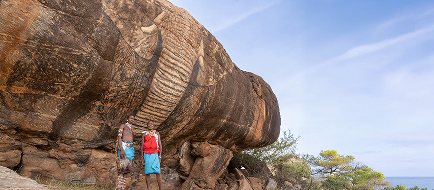 Two Samburu men in traditional attire stand beside a massive, elephant carving on a rock formation near Sarara Camp.