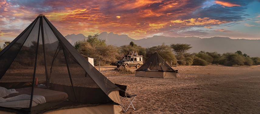 Canvas tents pitched on an open sandy riverbed at sunset, with a safari vehicle in the background and vivid evening sky.