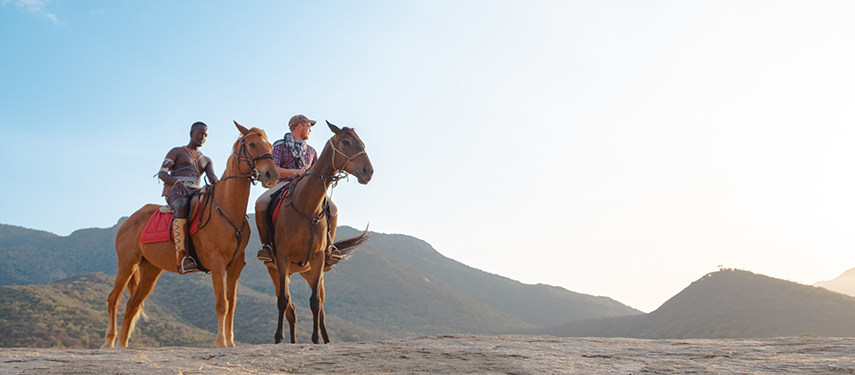 Two riders on horseback, one Samburu guide and one guest, take in the mountain scenery on a clear morning.