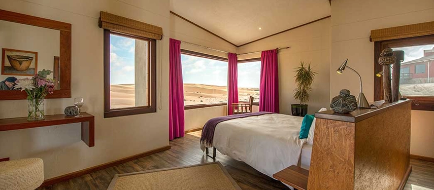 Bright, stylish bedroom at Desert Breeze Lodge with panoramic views of desert dunes through large corner windows and vibrant magenta curtains.