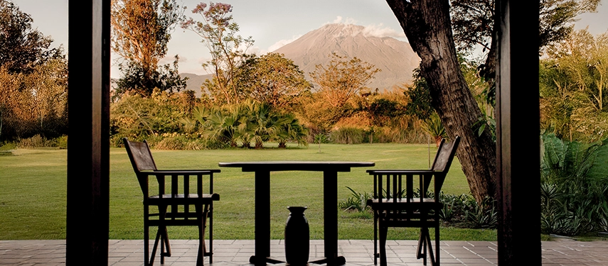 View from a private garden cottage veranda at Legendary Lodge in Arusha, with outdoor seating overlooking manicured lawns and Mount Meru rising in the distance.
