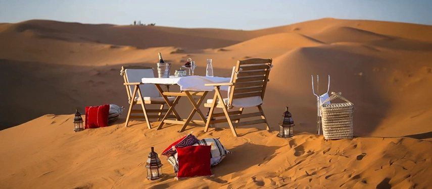 Private dining table set on desert dunes with lanterns, wine, and colourful cushions.
