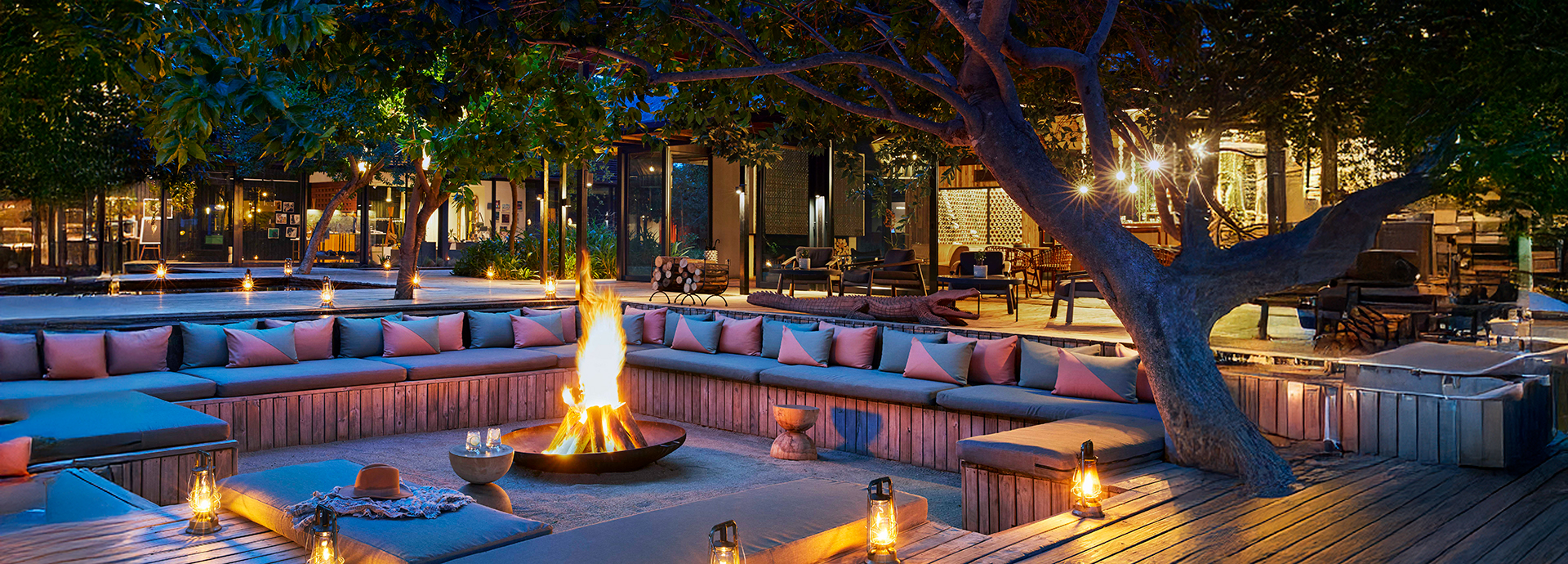 Lantern-lit fire deck at Lion Sands River Lodge with colourful cushions, ambient lighting, and a glowing central fire under tree canopy.